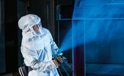 Eine Person in einem schützenden Anzug sprüht eine Flüssigkeit, während blauer Nebel aus der Sprühvorrichtung aufsteigt. Der Hintergrund ist dunkel, was den Fokus auf die Person und die Sprühaktion lenkt.